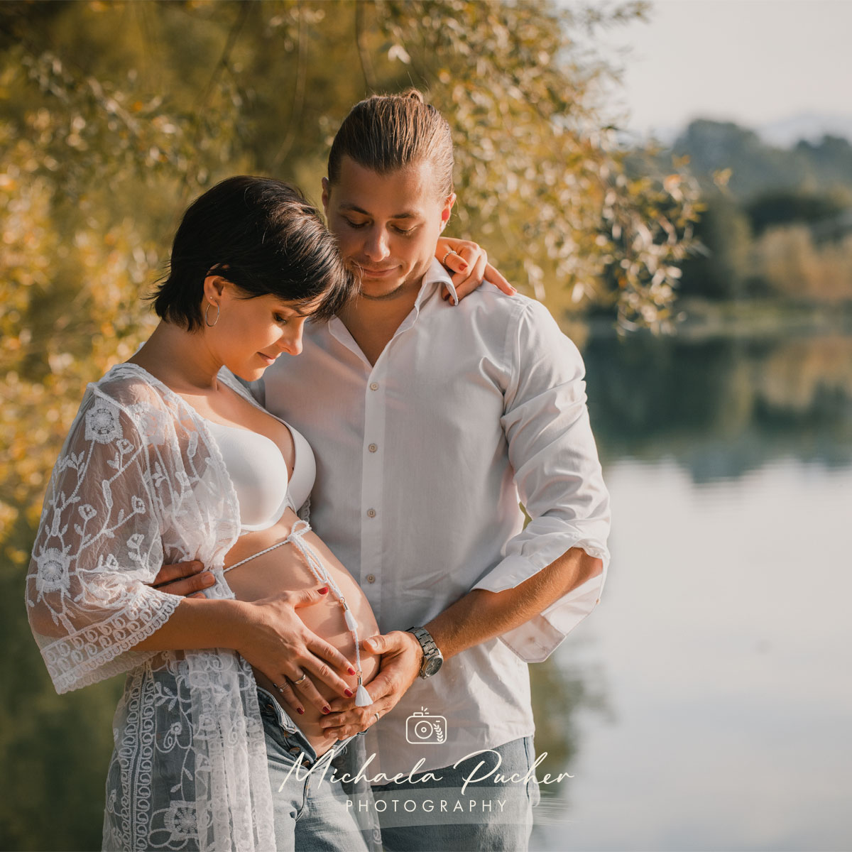 Schwangere steht mit ihrem Partner am See, beide halten liebevoll den Babybauch und schauen lächelnd darauf.